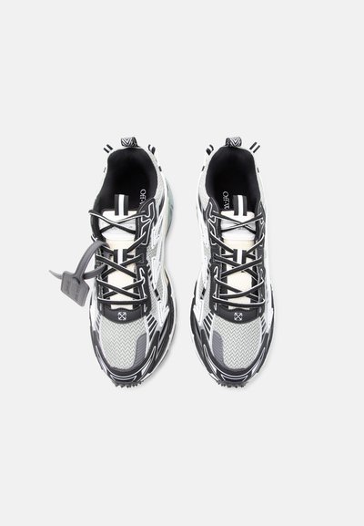 Ζευγάρι αθλητικών παπουτσιών σε μαύρο, άσπρο και γκρι χρώμα με ύφασμα από δίχτυ, διακοσμητικές λεπτομέρειες με μοτίβο, μαύρα κορδόνια και γκρι ταμπελάκι προσαρτημένο στο αριστερό παπούτσι.
