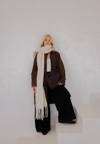 Bruine suède lange jas, crèmekleurige sjaal met franjes, wit topje, zwarte wijde broekspijpen en een donkerbruine clutch, staand op witte trappen.