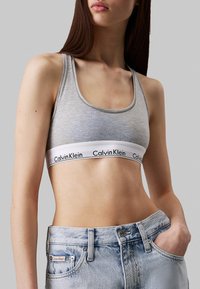 Sujetador deportivo de algodón gris con un escote redondo y una banda blanca ancha con el logotipo en negro de "Calvin Klein". Combinado con jeans azul desgastados.