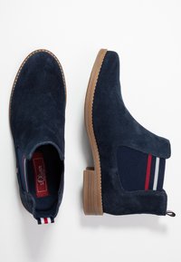 Botines de ante azul marino con paneles laterales elásticos que presentan rayas rojas y blancas, suela de goma y una lengüeta en la parte posterior para facilitar su uso.