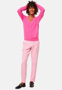 Cardigan rose tricoté avec des boutons devant et un col en V, associé à un pantalon rose clair. Le mannequin porte des chaussures noires avec des lanières.