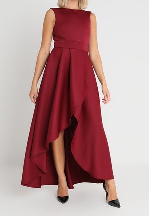 Mouwloze bordeaux jurk met een hoge-lage zoom, een getailleerde taille en een asymmetrische gelaagde rok, gedragen met zwarte puntenhakken.