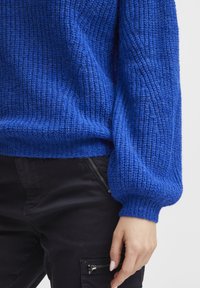 Maglione blu lavorato a maglia con finitura testurizzata, vestibilità ampia e maniche a palloncino. Abbinato a pantaloni neri con dettagli in cerniera.