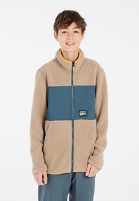 Chaqueta de forro polar con paneles beige y verde oscuro, cierre con cremallera en la parte delantera, cuello alto, bolsillo tipo canguro y parche de logo en el pecho. Textura suave.