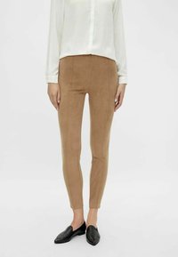 Beżowe legginsy zamszowe o dopasowanym kroju i ozdobnych szwach, zestawione z białą koszulą na guziki i czarnymi mokasynami. Gładka faktura.