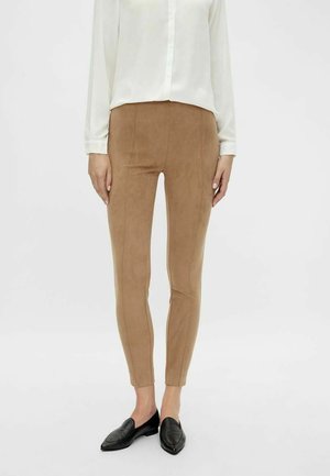 Tělově hnědé legíny z broušené kůže s úzkým střihem a švovými detaily, kombinované s bílou košilí na knoflíky a černými mokasíny. Hladká textura.