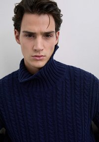 Pull en maille bleu marine avec un col haut à côtes et un motif en cable texturé, caractérisé par une coupe décontractée et des détails de couture proéminents.