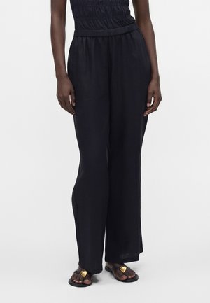 Personne portant un pantalon noir ample et des sandales noires avec des décorations dorées en forme de cœur sur fond blanc.