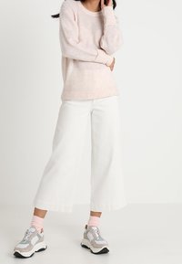 Frau in einem hellrosa Pullover, weißen, weit geschnittenen, cropped Hosen, rosa Socken und beigen Sneakern, die vor einem weißen Hintergrund steht.