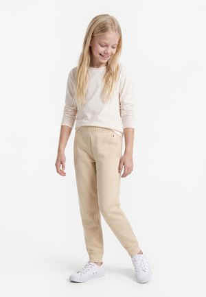 Jong meisje met lang blond haar, draagt een crèmekleurig shirt met lange mouwen, beige trainingsbroek en witte sneakers, staat en glimlacht.