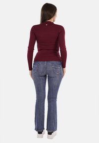 Top a maniche lunghe a costine bordeaux abbinato a jeans svasati blu. Dettagli notevoli includono un piccolo emblema sulla schiena e sneakers bianche.