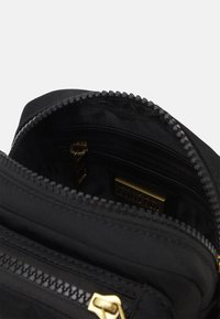 Sort lynlåset taske med åben hovedrum, der viser sort stofindvendigt og en guldfarvet lynlåstrækker i inderlommen.