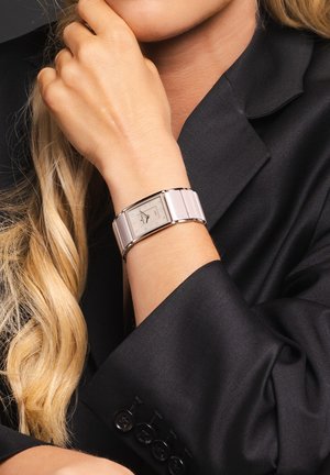 Silberne rechteckige Uhr mit grauem Zifferblatt und Diamantmarkierungen, ausgestattet mit einem glatten, hellrosefarbenen Metallarmband, getragen an einem Handgelenk in einem schwarzen Blazer.
