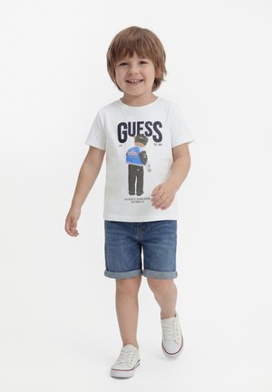 Smilende ung dreng går, iført en hvid grafisk t-shirt med teksten "GUESS", optrukne cowboyshorts og hvide sneakers på en ensfarvet baggrund.