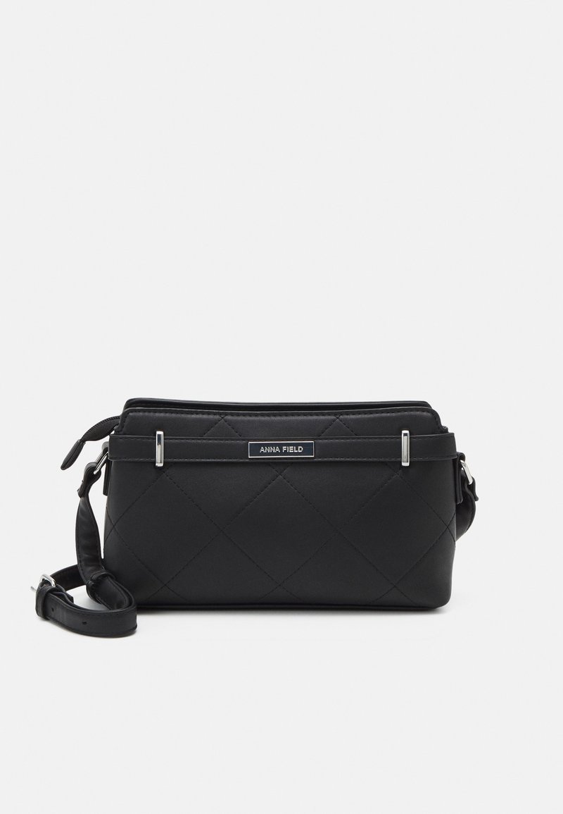 Sac bandoulière matelassé noir avec sangle ajustable et quincaillerie argentée, comportant une petite étiquette rectangulaire "ANNA FIELD" à l'avant.