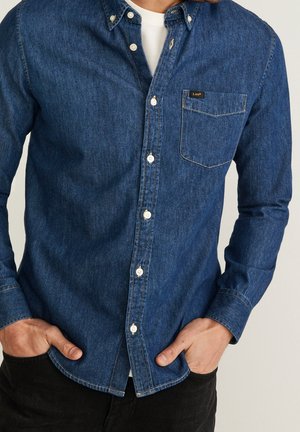 Mand iført en blå denimskjorte med knapper og en brystlomme med mærket "Lee", hvid undertrøje, hænder i lommerne på sorte bukser.