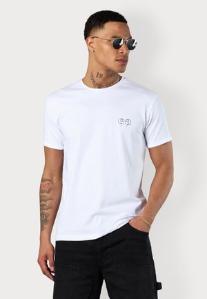 White cotton t-shirt with a round neckline and short sleeves. Features a small black logo on the chest and is paired with black pants.