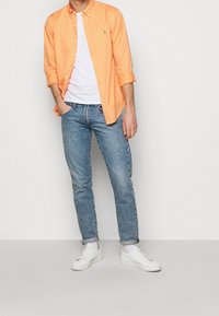 Oranžová košile na knoflíky přes bílý tričko, v kombinaci se světle modrými džíny a bílými teniskami. Košile má na hrudi logo.