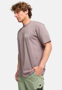 Paarse katoenen t-shirt met een klein logo op de voorkant. Korte mouwen en een ronde hals. Gecombineerd met lichtgroene broeken.