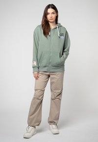 Groene ritssluiting hoodie met voorzakken, trekkoord capuchon en grafische print. Gepaard met beige broek en witte sneakers. Glad stofoppervlak.