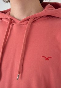 Korallefarbene Kapuzenjacke mit einer vorderen Tasche, weicher Textur, verstellbaren Kordeln und einem kleinen roten gestickten Logo auf der Brust.