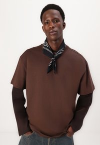 Braunes Kurzarm-T-Shirt, das über einem langärmeligen schwarzen Oberteil getragen wird, mit einem schwarzen Bandana um den Hals, vor einem neutralen Hintergrund präsentiert.