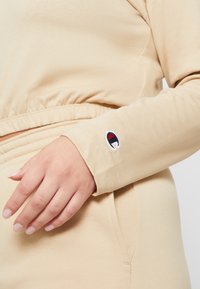 Die Hand einer Frau ruht in der Nähe der Taille und trägt ein beigefarbenes Langarmoberteil sowie eine Hose mit kleinem Champion-Logo am Ärmelbund.