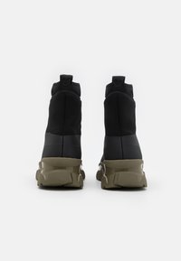 Zapatillas negras de caña alta con un acabado mate, suela de goma texturizada en verde oliva y tiradores en la parte posterior para ponérselas fácilmente.