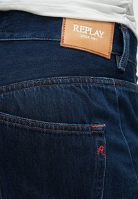 Σκούρο μπλε τζιν με πορτοκαλί ραφές, δερμάτινη ετικέτα με το κείμενο "REPLAY" και τσέπη με μικρή κόκκινη λεπτομέρεια «R».