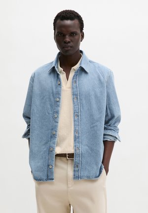 Jeune homme portant une veste en jean bleu clair sur une chemise beige et un pantalon beige, debout les mains dans les poches devant un fond uni.