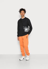 Μαύρο μπλουζάκι με μακριά μανίκια, πορτοκαλί φόρμες, άσπρα αθλητικά παπούτσια Nike και μια μαύρη και γκρι τσάντα ώμου. Απλός σχεδιασμός, άνετη εφαρμογή και ορατή σήμανση.