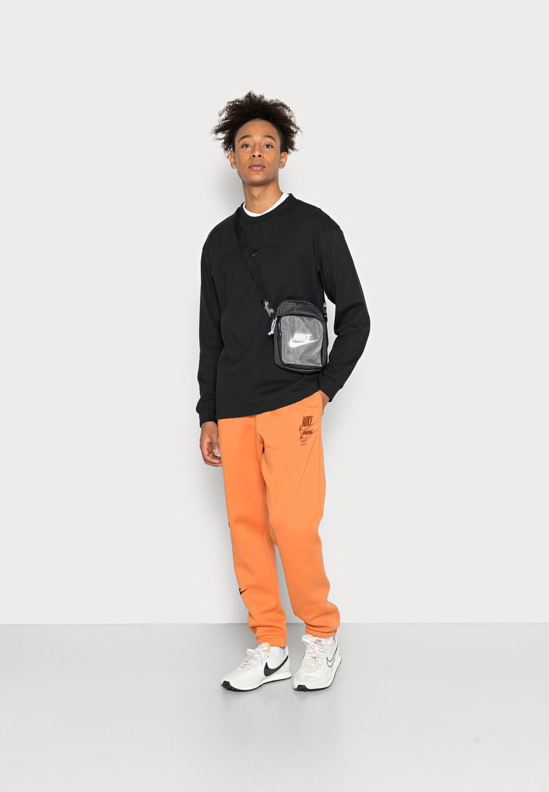 Schwarzes Langarmshirt, orange Sweatpants, weiße Nike-Sneaker und eine schwarz-graue Umhängetasche. Einfaches Design, lässige Passform und sichtbare Markenkennzeichnung.