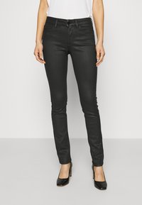 Jeans ajustados negros con un diseño entallado, hechos de tela vaquera, que presentan una cintura alta y un cierre de botón. Llevados con tacones negros.