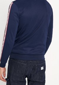 Navy blue sweatshirt with a ribbed collar and hem, features red and white striped side accents labeled "TOMMY JEANS." Worn with pinstripe pants.