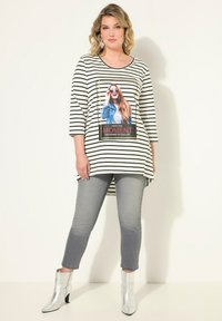 Gestreiftes, übergroßes Oberteil mit einem grafischen Druck, der eine Frau zeigt, kombiniert mit grauen Jeans und metallischen Ankle Boots. Dreiviertelärmel.