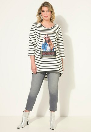 Gestreiftes, übergroßes Oberteil mit einem grafischen Druck, der eine Frau zeigt, kombiniert mit grauen Jeans und metallischen Ankle Boots. Dreiviertelärmel.