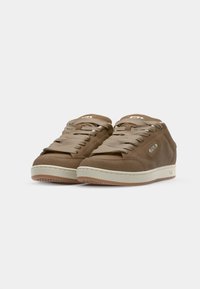 Baskets basses en daim marron avec une semelle beige clair, dotées de lacets ronds et d'un petit logo sur le côté. Surface texturée et design à bout arrondi.