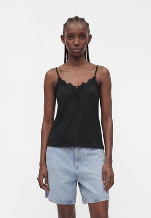 Donna con capelli intrecciati che indossa una canottiera nera con rifiniture in pizzo e pantaloncini di denim azzurri, in piedi davanti a uno sfondo chiaro e uniforme.