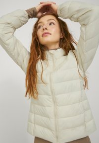 Lichtbeige pufferjack met een rits aan de voorkant, met een gestikt ontwerp en hoge kraag. Zachte textuur en iets uitlopende zoom.