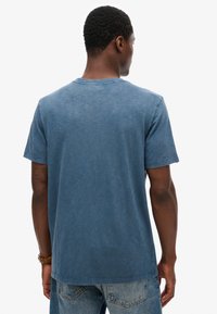 Blå bomulds T-shirt med korte ærmer og rund halsudskæring. Har en falmet tekstur og en lige kant. Bæres med lyse jeans.