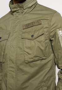 Olivgrön jacka med hög krage, dragkedja och tryckknappsstängning, två framfickor med klaff och en sidodragkedjeficka, med en tygetikett på bröstet.