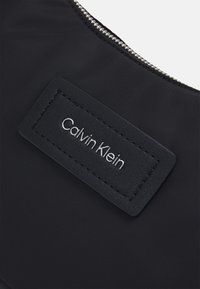Musta kangas kott, millel on tekstuuriga nahasilt, millel on reljeefne "Calvin Klein" logo ja hõbedane lukustus ülemises servas.