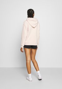 Person med flätat hår som bär en ljusrosa hoodie, svarta jeansshorts, vita strumpor och vita högskor, som står framför en neutral bakgrund.