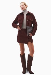 Bordowy sztruksowy żakiet z dwiema kieszeniami na piersi, zestawiony z pasującą spódnicą. Szary golf i brązowe botki na wysokim obcasie do kolan. Ciemna torebka.