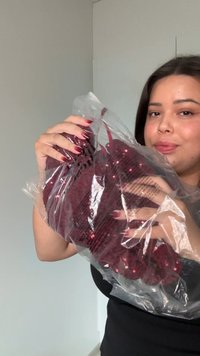 Tamsiais plaukais moteris su raudonais nagų lakais laiko skaidrų plastikinį maišelį, kuriame yra bordo spalvos pačių sekančių drabužis, priešais šviesią, paprastą sieną.