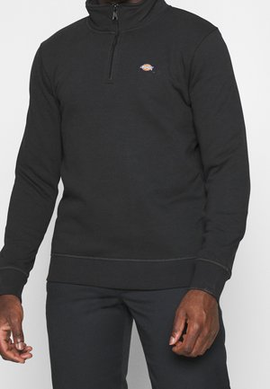 Hombre vistiendo un sudadera negra con media cremallera y un pequeño logotipo redondo multicolor en el lado izquierdo del pecho, junto con pantalones negros a juego.