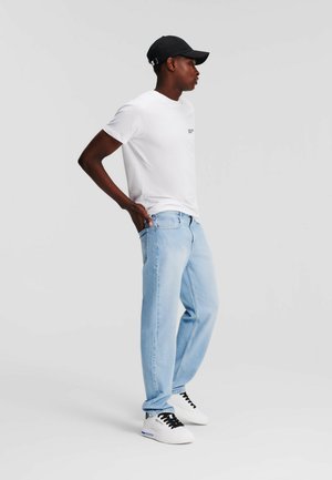 Bela majica, svetlo modri jeans in črna kapa. Model stoji obrnjen na stran, roke na bokih, oblečen v bele športne copate s črnimi detajli.