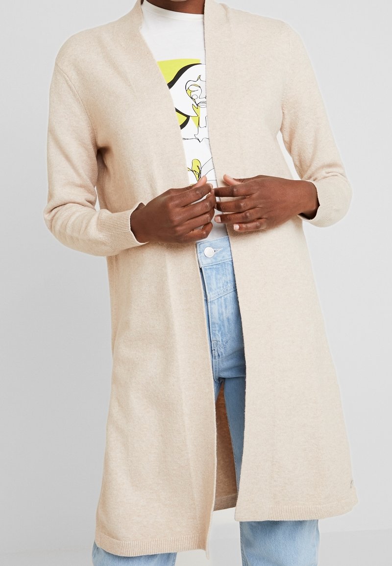 Beige knitted cardigan with an open front, long sleeves, and ribbed cuffs. Worn over a white shirt with a graphic design.