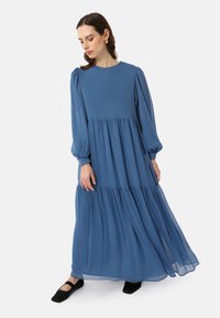 Blue long-sleeve maxi dress with gathered tiers, round neckline, and balloon sleeves, made of lightweight fabric. Worn with black flats.