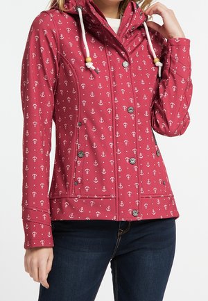 Femme portant une veste rouge avec un motif d'ancre blanche, des cordons blancs avec des perles en bois et un jean bleu, main touchant le col de la veste.